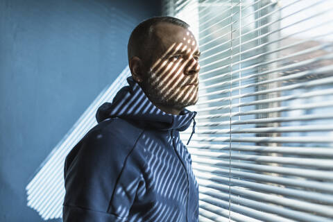 Pensive man looking out of venetian blind window stock photo