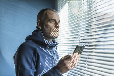 Nachdenklicher Mann mit Smartphone und Kopfhörern, der aus einem Jalousienfenster schaut - MCVF00279