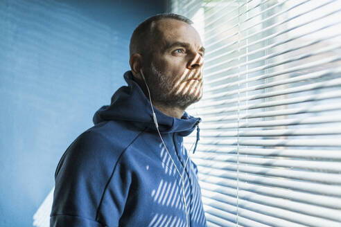 Nachdenklicher Mann mit Ohrstöpseln, der aus einem Jalousienfenster schaut - MCVF00280