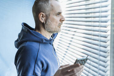 Nachdenklicher Mann mit Smartphone und Kopfhörern, der aus einem Jalousienfenster schaut - MCVF00282