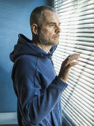 Nachdenklicher Mann mit Ohrstöpseln, der aus einem Jalousienfenster schaut - MCVF00284