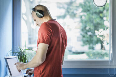 Mann mit Kopfhörern, der ein digitales Tablet in der Nähe eines Fensters zu Hause benutzt - MCVF00286