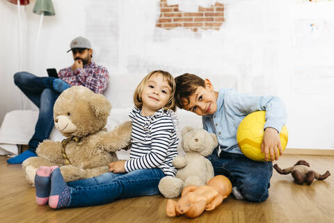 Brother and sister sitting in playroom with cuddly toys, father in background stock photo