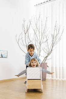 Brother pushing sister, sitting in a doll buggy, playing at home - JRFF04333