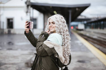 Portrait of smiling young woman taking selfie with smartphone on platform - LJF01495