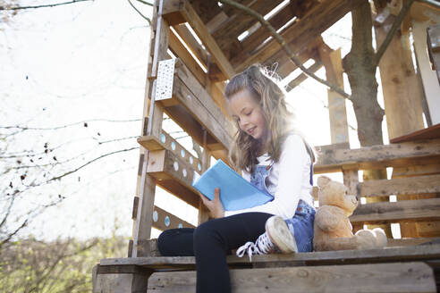 Mädchen sitzt im Garten am Baumhaus und schreibt etwas in ihr blaues Tagebuch - HMEF00883