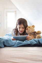 Porträt eines lächelnden Mädchens, das mit einem Teddybären auf dem Bett liegt und ein digitales Tablet benutzt - LVF08778