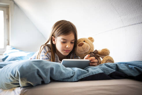 Porträt eines Mädchens, das mit einem Teddybär auf dem Bett liegt und ein digitales Tablet benutzt - LVF08779