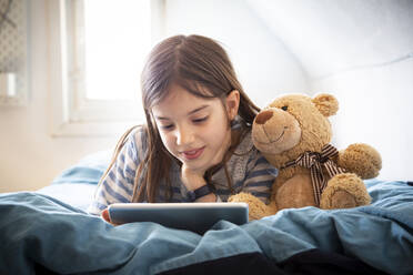 Porträt eines lächelnden Mädchens, das mit einem Teddybären auf dem Bett liegt und ein digitales Tablet benutzt - LVF08780