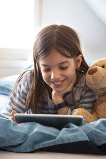 Porträt eines lächelnden Mädchens, das mit einem Teddybären auf dem Bett liegt und ein digitales Tablet benutzt - LVF08781