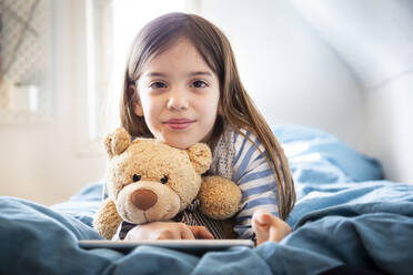 Porträt eines lächelnden Mädchens auf dem Bett liegend mit Teddybär und digitalem Tablet - LVF08782