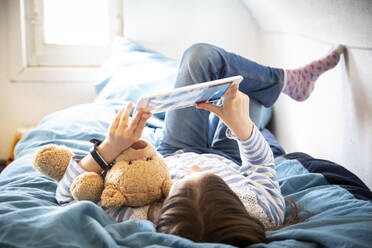Little girl lying on bed with teddy bear using digital tablet - LVF08783