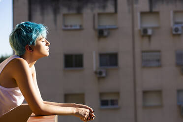 Androgyne junge Frau mit blauem Haar genießt das Sonnenlicht auf dem Balkon - ERRF03428
