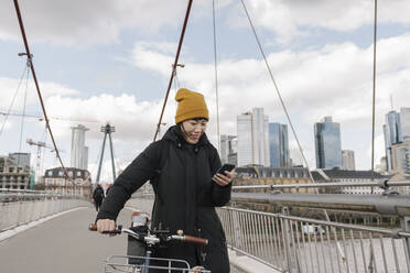 Smiling woman with bicycle using smartphone on a bridge, Frankfurt, Germany - AHSF02232