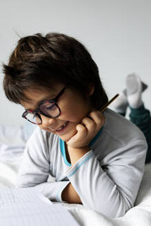 Porträt eines kleinen Jungen, der auf dem Bett liegt und Hausaufgaben macht - VABF02770