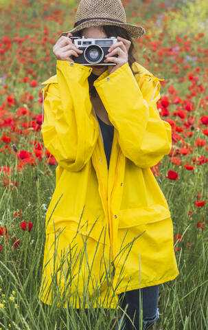 Junge Frau in gelbem Regenmantel beim Fotografieren auf einer Blumenwiese mit Mohnblumen, lizenzfreies Stockfoto