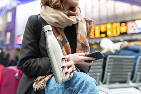 Woman sitting at train station with thermos flask and mobile phone - WPEF02799