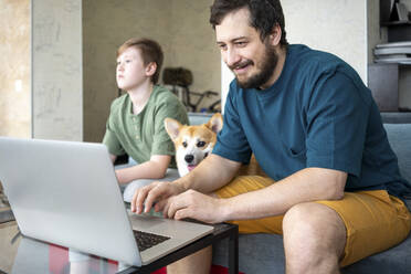 Porträt eines lächelnden Vaters, der zu Hause einen Laptop benutzt, während sich sein Sohn im Hintergrund langweilt - VPIF02300