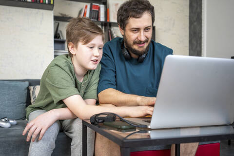 Porträt von Vater und Sohn, die zusammen auf der Couch zu Hause sitzen und einen Laptop benutzen, lizenzfreies Stockfoto