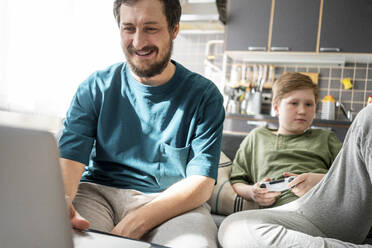 Porträt eines lächelnden Vaters, der zu Hause einen Laptop benutzt, während sich sein Sohn im Hintergrund langweilt - VPIF02305