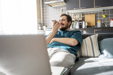 Portrait of smiling man sitting on couch at home using mobile phone - VPIF02308