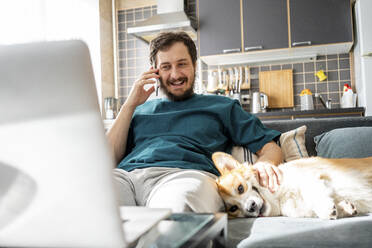Porträt eines lächelnden Mannes am Telefon mit Hund auf der Couch zu Hause - VPIF02309