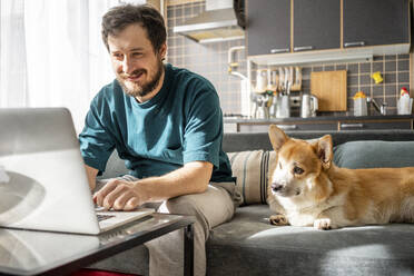 Porträt eines lächelnden Mannes, der zu Hause auf der Couch sitzt und einen Laptop benutzt - VPIF02310