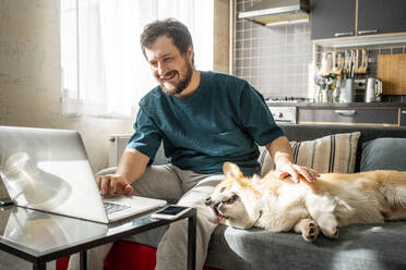 Porträt eines lächelnden Mannes, der zu Hause auf der Couch sitzt und einen Laptop benutzt - VPIF02311