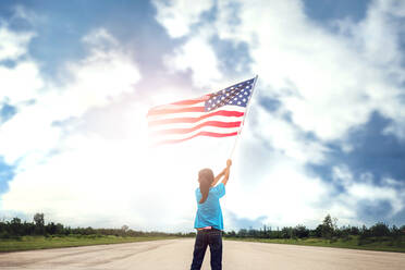 Rückansicht des Mädchens, das die amerikanische Flagge hält, während es auf der Straße gegen den Himmel steht - EYF03843