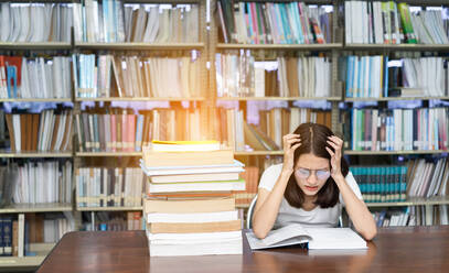 Junge Frau mit Kopf in den Händen studiert am Schreibtisch in der Bibliothek - EYF03925