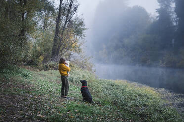 A woman is holding a baby and looking at a dog near a river - CAVF79046