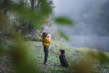 A woman is holding a baby and looking at a dog near a river - CAVF79047