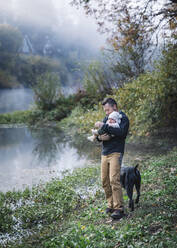 Ein Mann hält ein Baby in der Nähe eines Flusses und einen Hund - CAVF79049