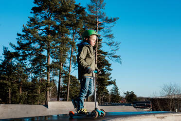 Boy standing on his scooter at a skatepark in the sunshine - CAVF79065