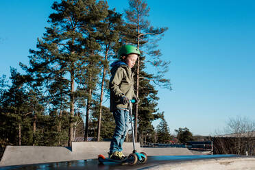 Boy standing on his scooter in a skatepark in the sunshine - CAVF79066