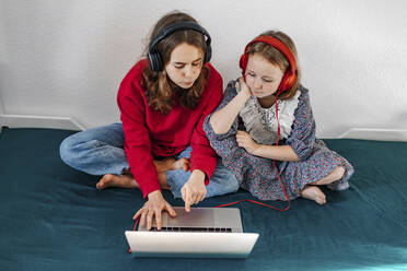 Portrait of two sisters with headphones sitting on bed looking at laptop - OGF00268