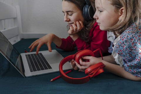 Zwei Schwestern mit Kopfhörern liegen auf dem Bett und benutzen einen Laptop, lizenzfreies Stockfoto