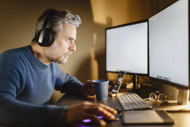 Älterer Mann mit Kopfhörern sitzt am Schreibtisch zu Hause und arbeitet am Computer - MCVF00295