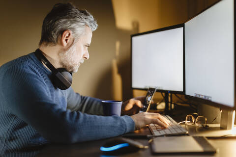 Älterer Mann sitzt am Schreibtisch zu Hause und arbeitet mit Smartphone und Computer, lizenzfreies Stockfoto