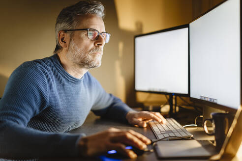 Älterer Mann sitzt am Schreibtisch zu Hause und arbeitet am Computer - MCVF00297