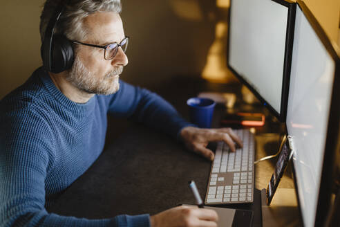 Mature man with headphones sitting at desk at home working with graphics tablet and computer - MCVF00300