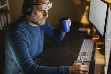 Reifer Mann mit Kopfhörern sitzt am Schreibtisch zu Hause und arbeitet mit Grafiktablett und Computer - MCVF00302
