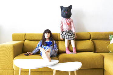 Two bored girls sitting on couch, watching TV, one wearing cat mask - ERRF03436
