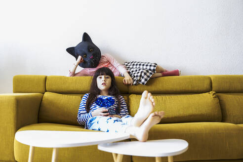 Zwei gelangweilte Mädchen sitzen auf der Couch und sehen fern, eine trägt eine Katzenmaske - ERRF03437