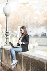 Lächelnde Frau mit Kaffee zum Mitnehmen, die an einer Wand sitzt und auf einen Laptop schaut - LJF01501