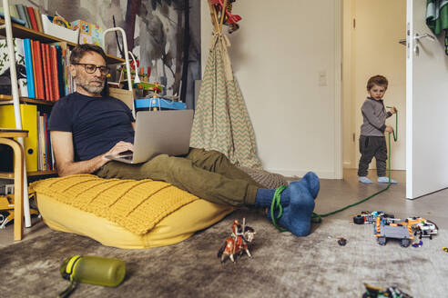 Son tying his father working on laptop in children's room - MFF05575