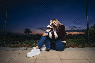 Young woman wearing fur jacket, sitting on the ground at night - MPPF00768