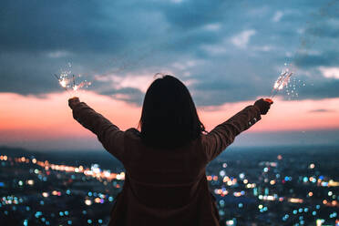 Rear View Of Woman Holding Sparklers Against Illuminated Cityscape At Dusk - EYF04279