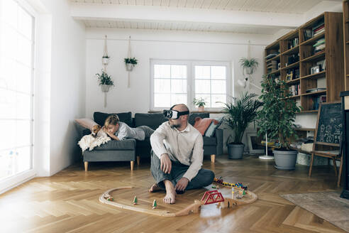 Vater sitzt in der Mitte von Spielzeug und benutzt eine VR-Brille, während der Sohn auf der Couch liegt - KMKF01295