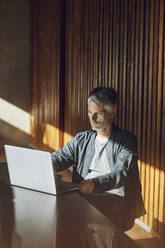 Lässiger Geschäftsmann, der in seinem nachhaltigen Büro sitzt und einen Laptop benutzt - MCF00635